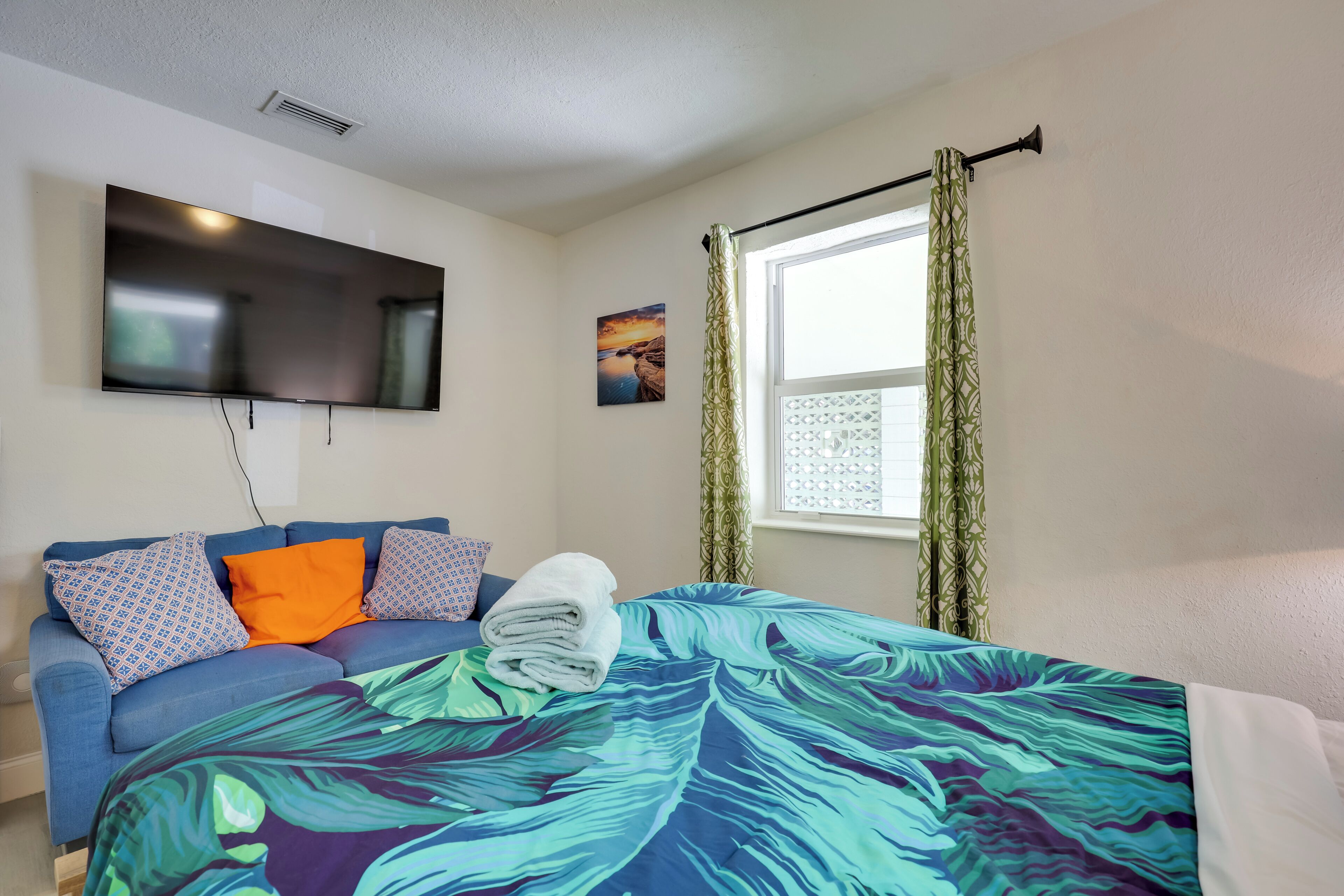 A bedroom with a bed, a flat-screen TV mounted on the wall, a window with curtains, and a sofa with patterned cushions.