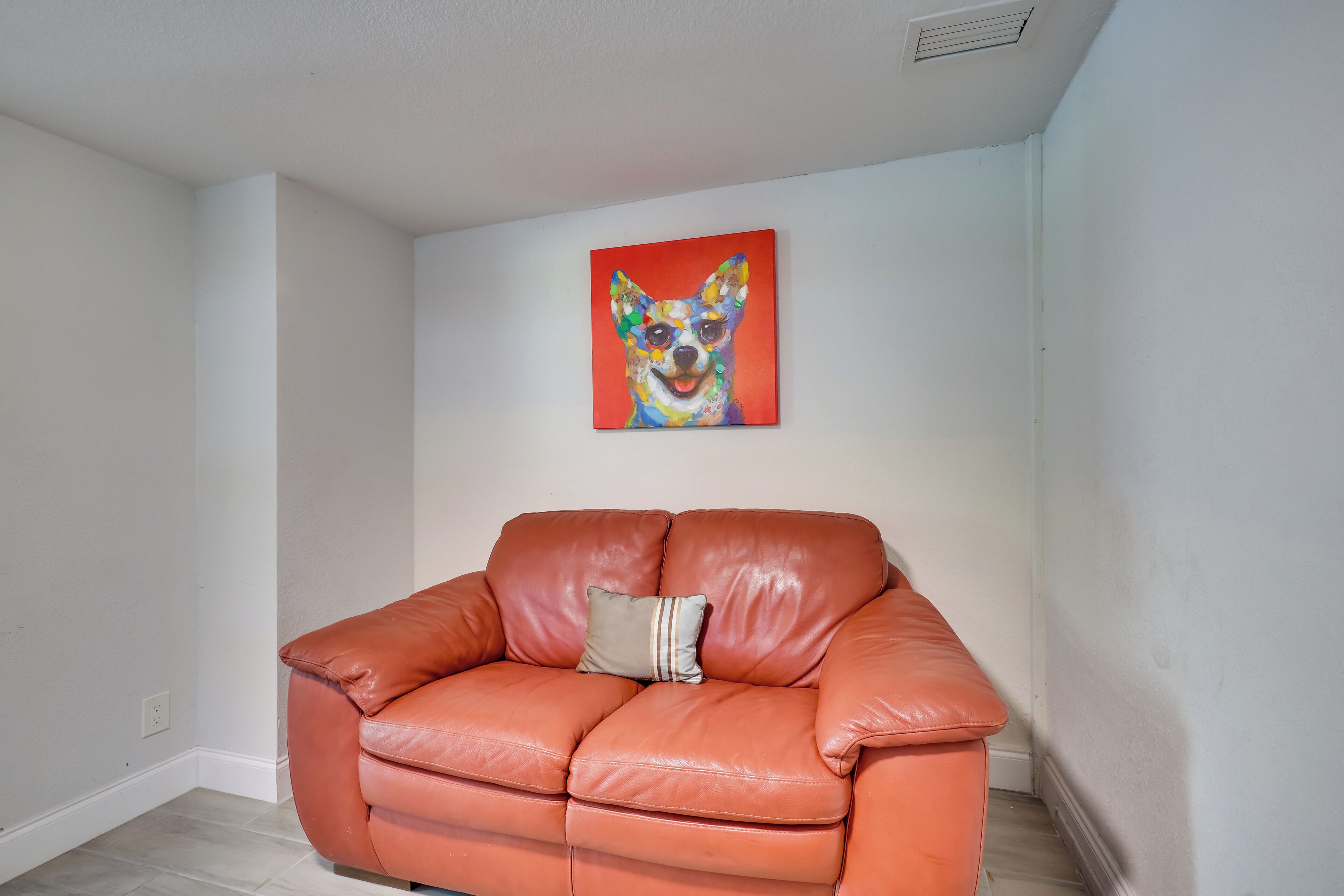 Una habitación con un sofá de cuero marrón, un cojín y un cuadro de un perro colorido en la pared.