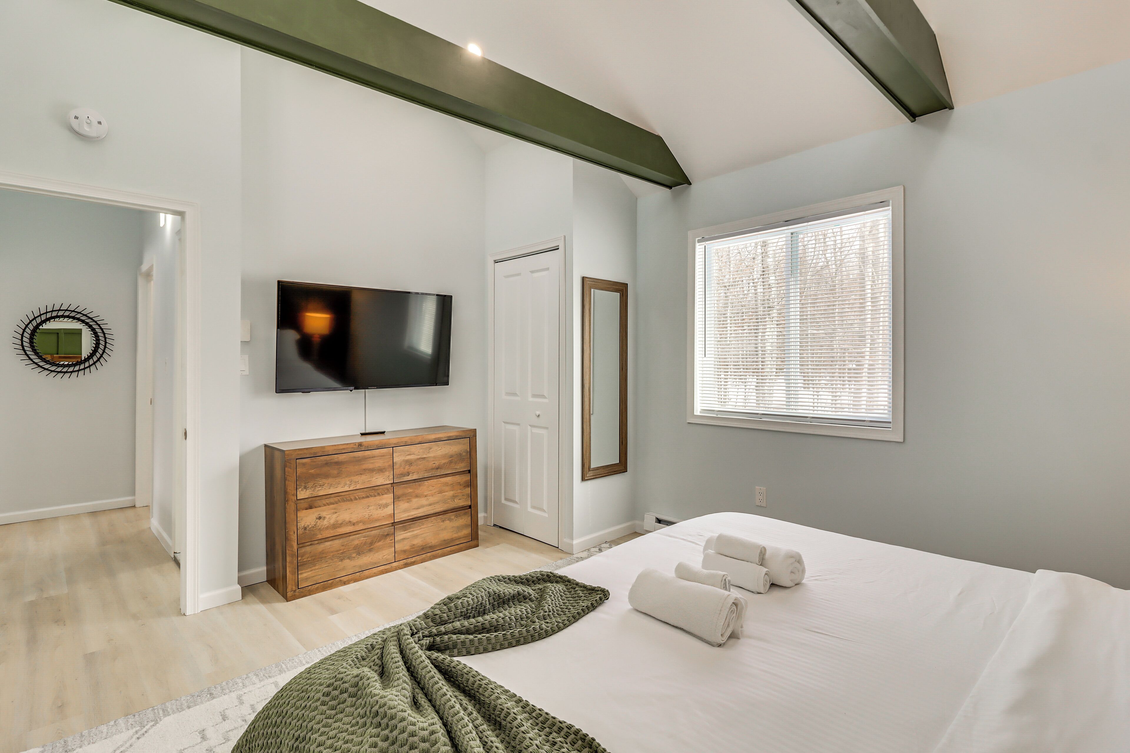 Un dormitorio con cama, televisor, cómoda, espejo y ventana con persianas.
