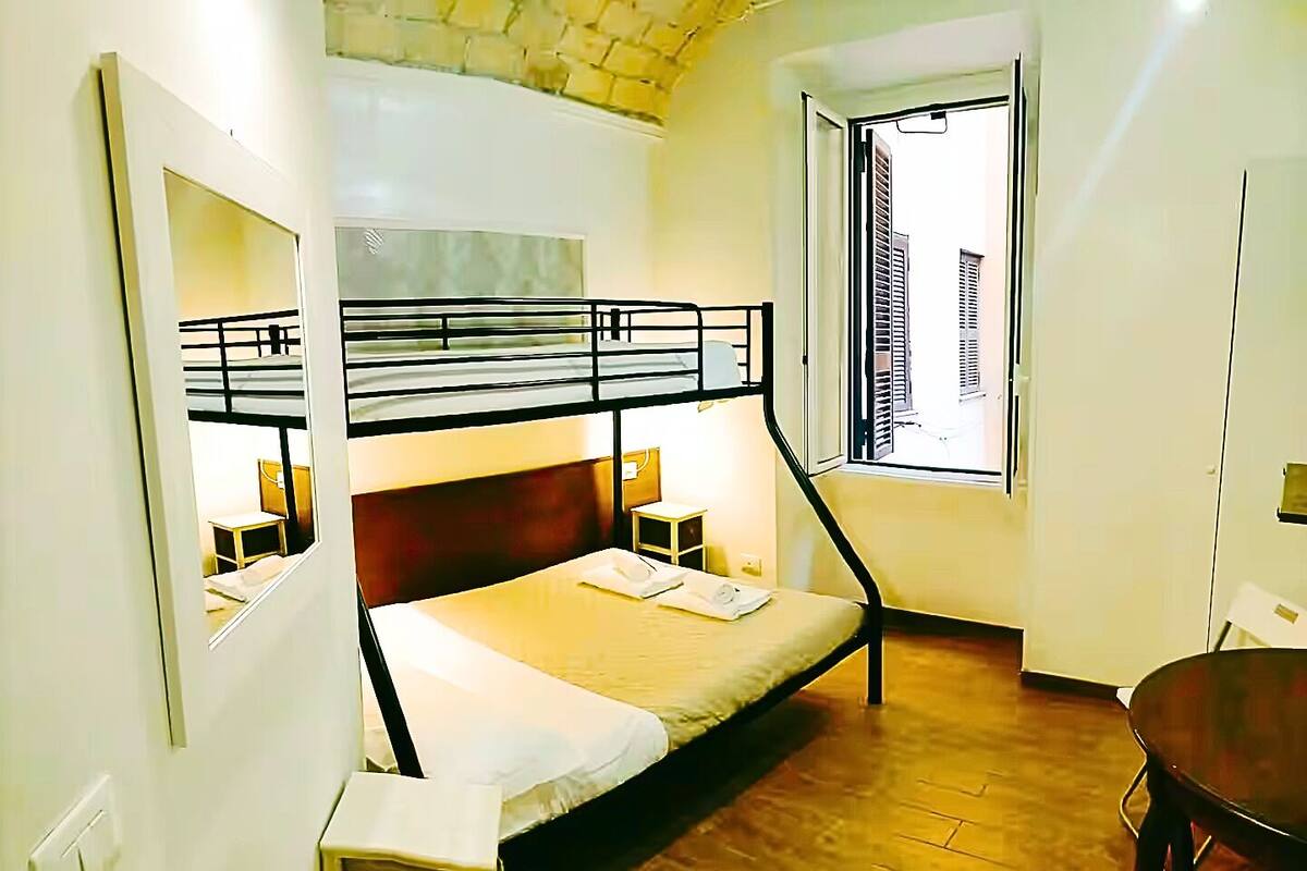 A room with bunk beds, a desk, a chair, a window with a view of buildings, and a wooden floor.