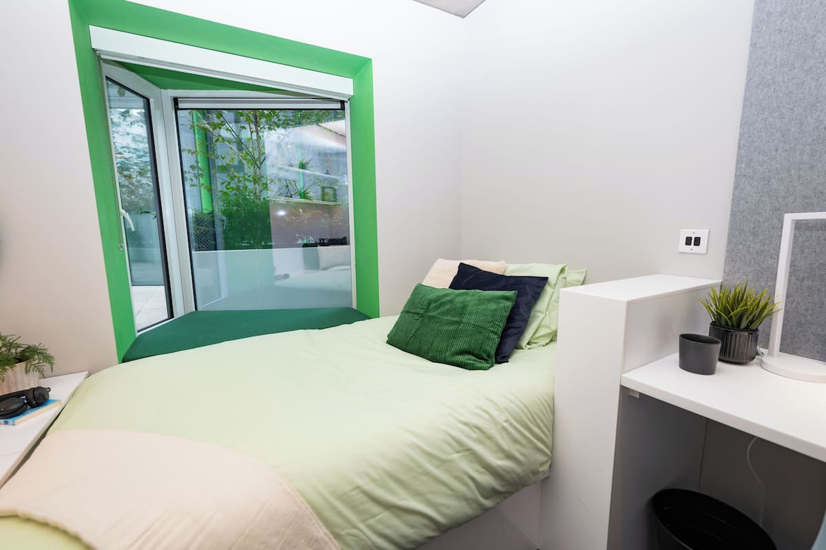 A modern bedroom with a green-framed window, a bed with green and dark green pillows, a desk with a plant, and a small trash bin.