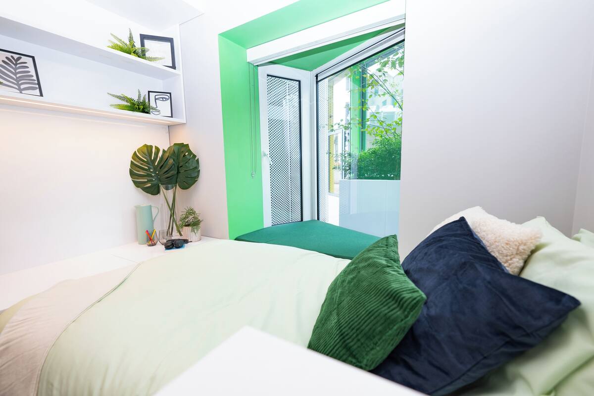 A modern bedroom with a bed, green and white pillows, a green plant, a small table with a plant and a mug, and a window with a view of greenery.