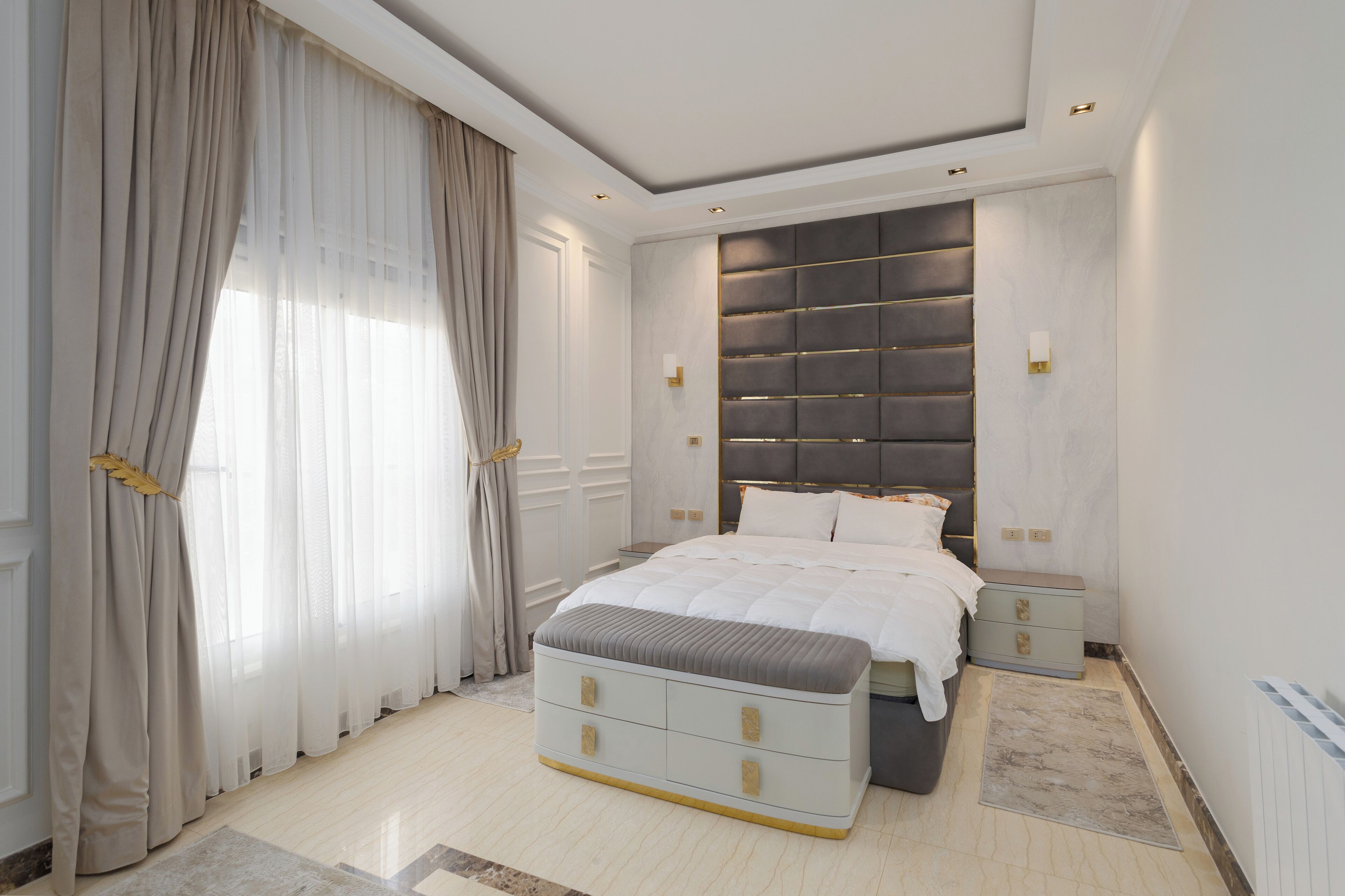 Une chambre à coucher avec un lit, des tables de chevet, une commode, un banc et une fenêtre avec des rideaux.