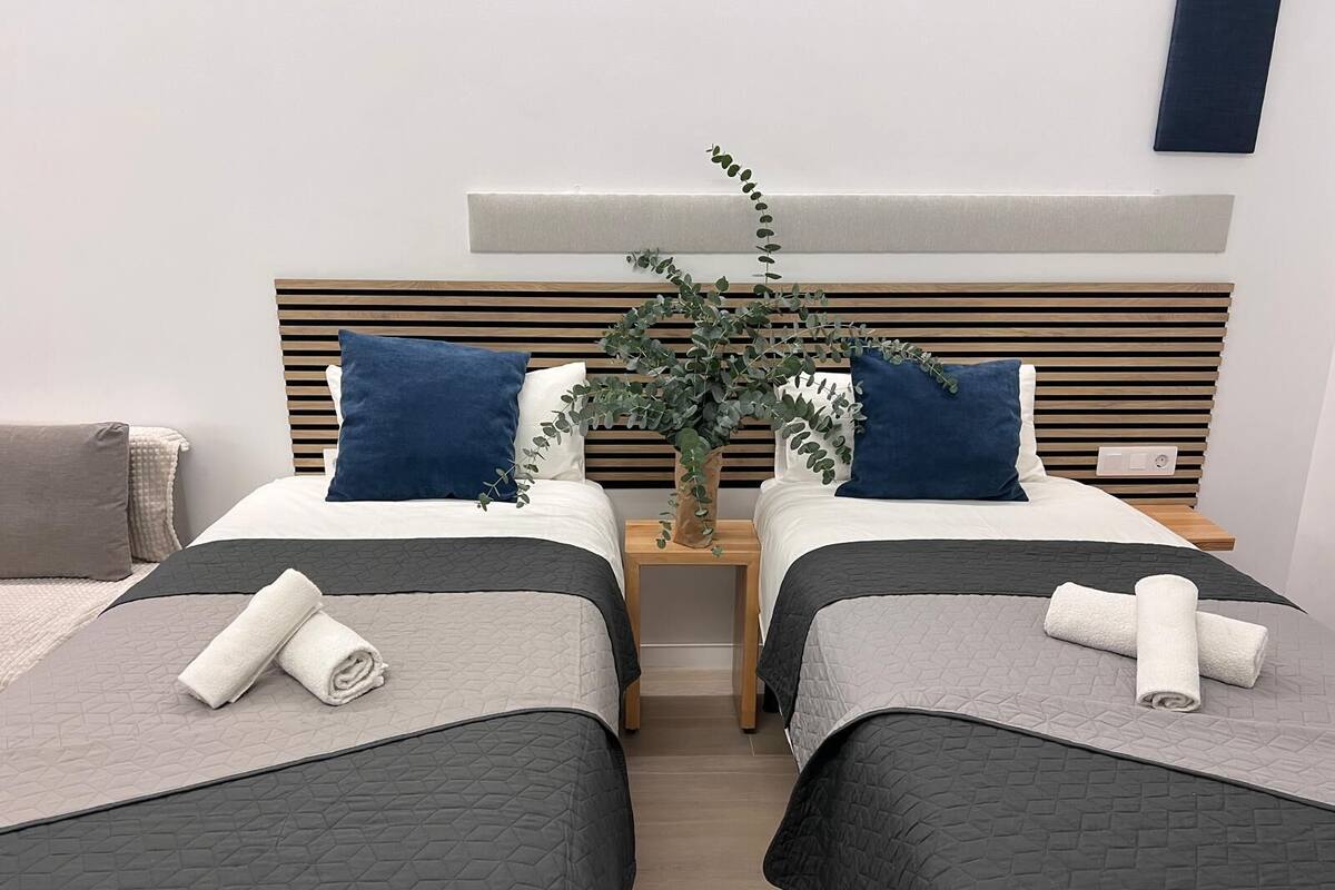 A hotel room with two beds, each with a grey quilted bedspread and a rolled white towel. There is a wooden headboard with horizontal slats and a small plant on the nightstand.