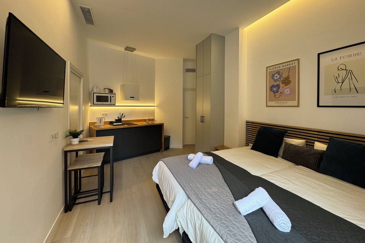 A modern hotel room with a bed, bedside table, and a small kitchenette.