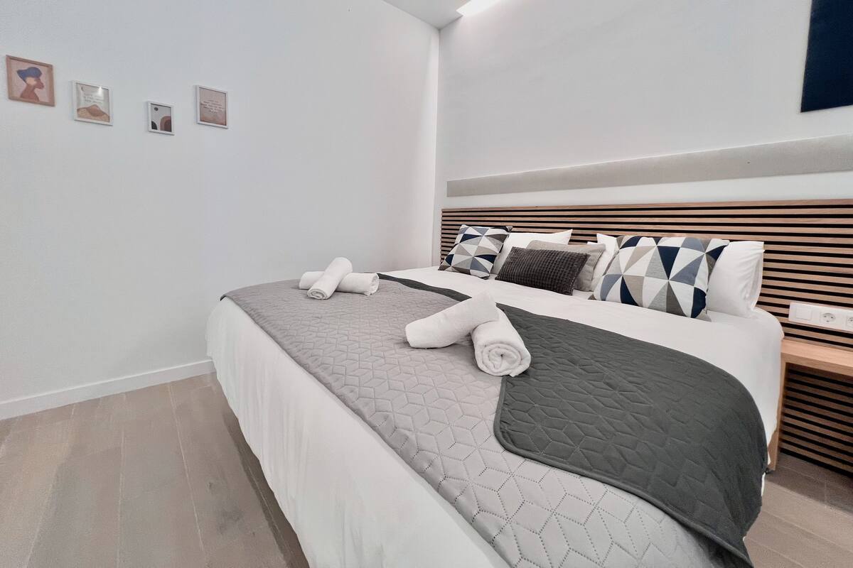 A modern bedroom with a neatly made bed, a headboard with geometric patterned pillows, and a wall with framed artwork.
