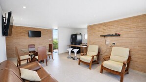 Un salon moderne avec un canapé en cuir, des chaises en bois, une table à manger en verre et un coin cuisine doté d’un mur en briques et d’un téléviseur fixé au-dessus.