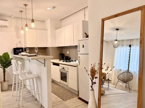 A modern kitchen with white cabinets, a built-in oven, and a coffee machine. There is a dining area with white chairs and a small table. - La Bohème Signature Apartment in La Marsa (La Marsa)