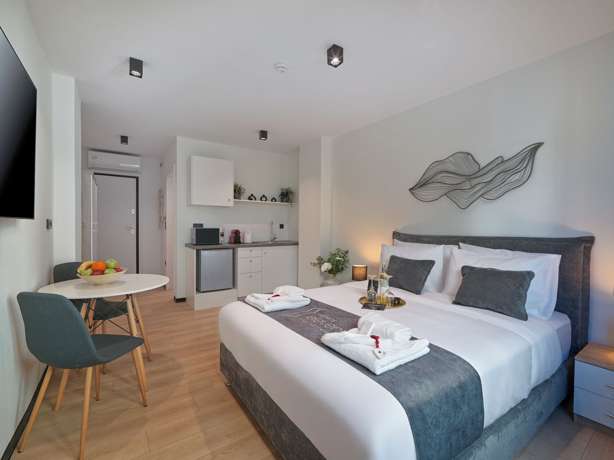 Une chambre d’hôtel moderne équipée d’un lit, d’une table de chevet, d’un petit bureau avec une chaise, d’une kitchenette et d’un téléviseur.