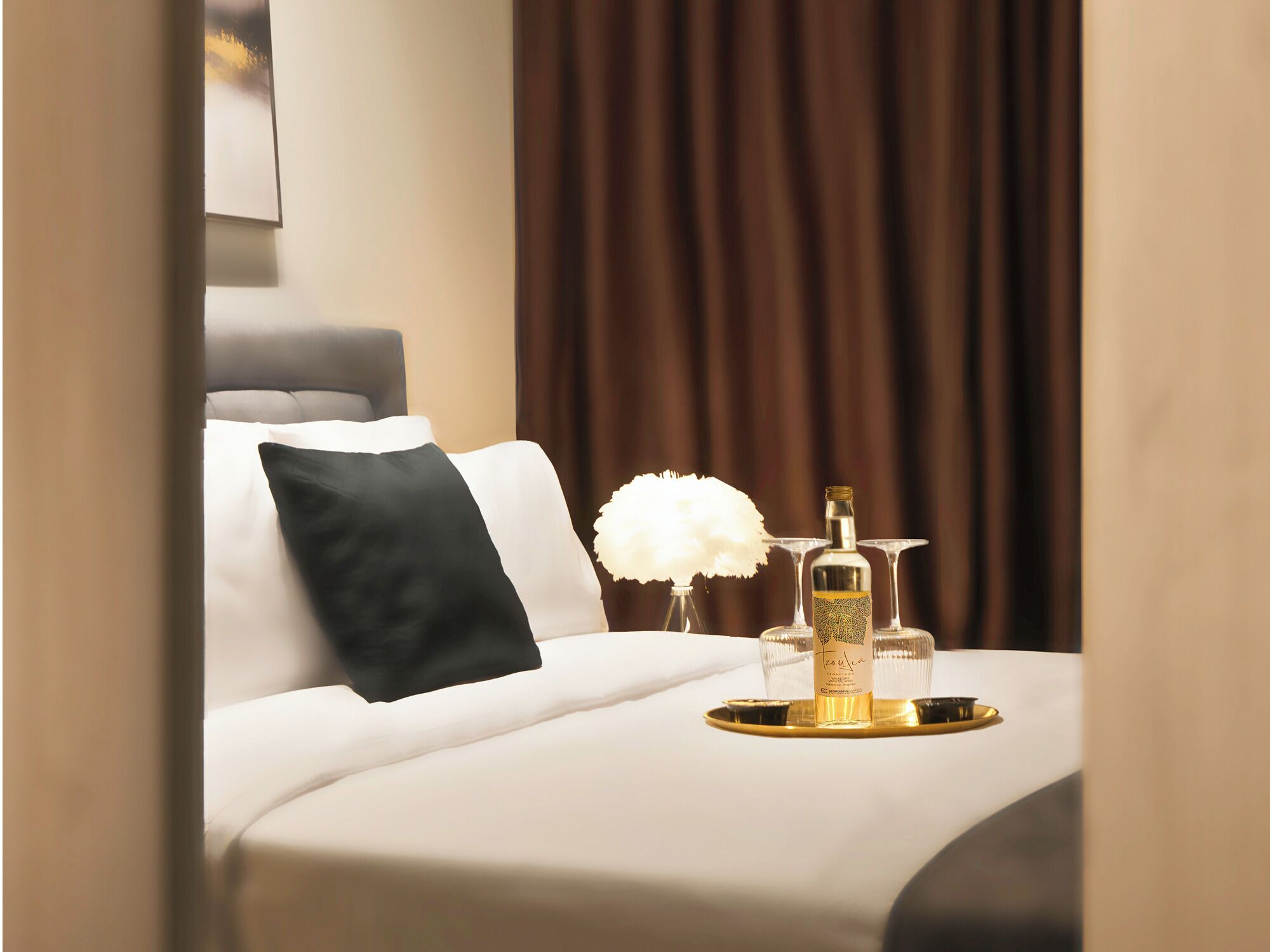 A neatly made bed with white and black pillows, a decorative white feather, a bottle, and glasses on a tray.