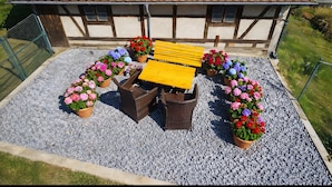Un toit recouvert de panneaux solaires, un bâtiment en bois, une terrasse en gravier, une table jaune, des chaises en osier et des pots de fleurs.