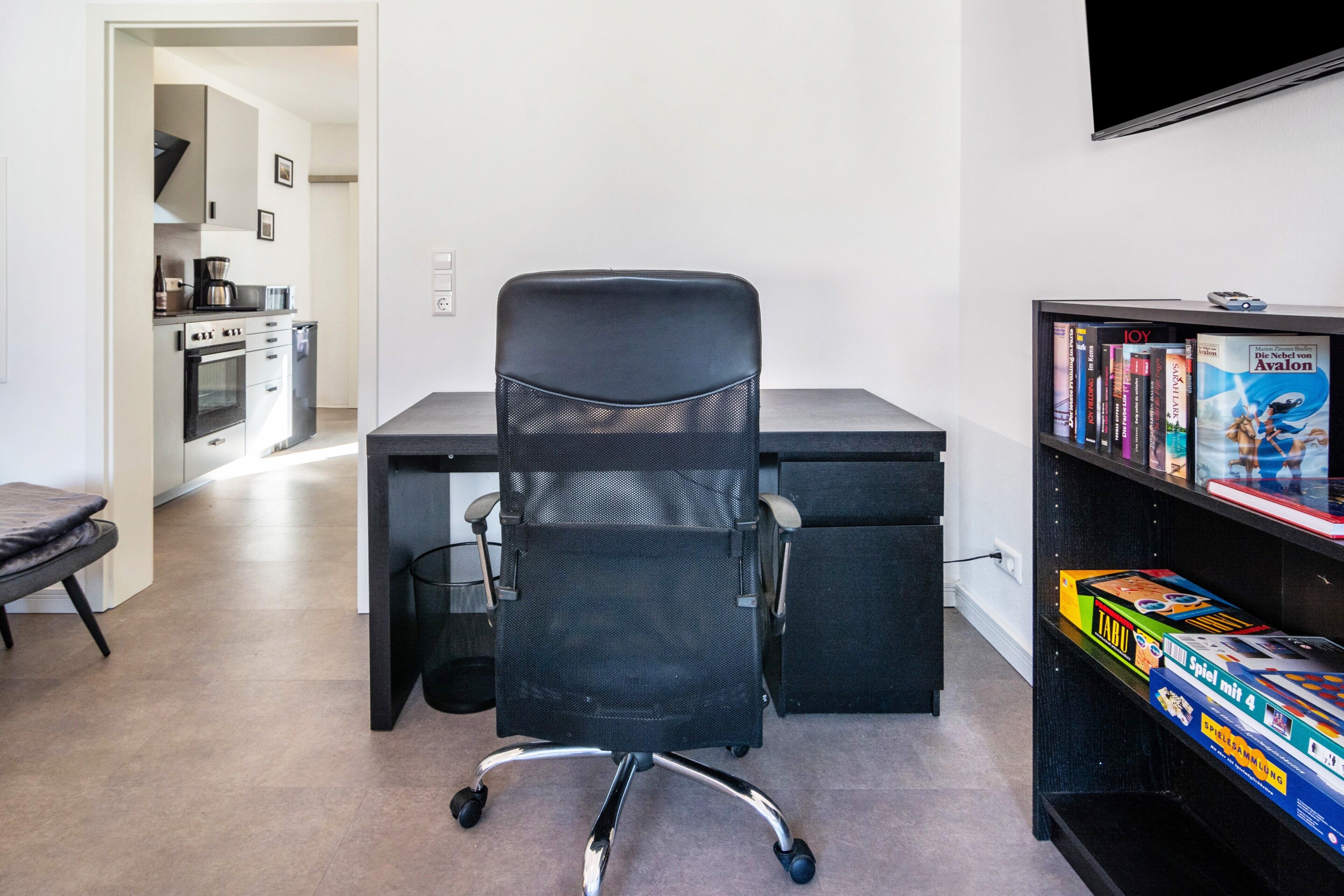 Un espace de bureau moderne comprenant un bureau, un fauteuil de bureau, une bibliothèque et, au fond, un coin cuisine.