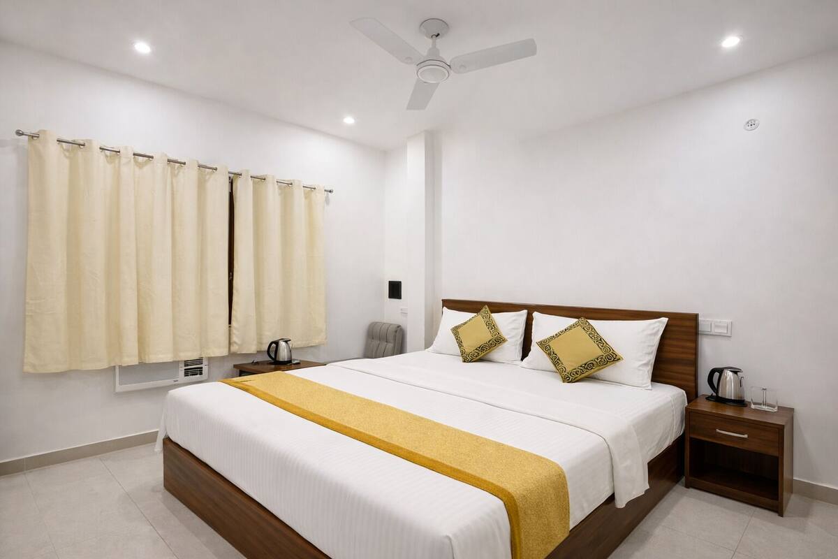 A hotel room with a large bed, a nightstand, a ceiling fan, a window with curtains, and a wall-mounted air conditioning unit.
