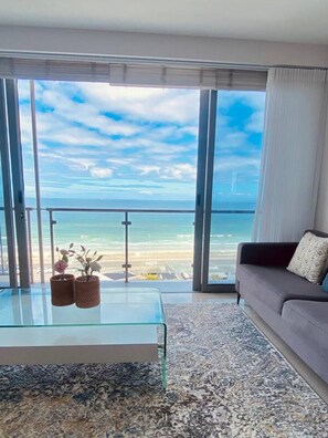 Ein modernes Wohnzimmer mit einem Glastisch, einem grauen Sofa und Blick auf das Meer durch große Fenster.