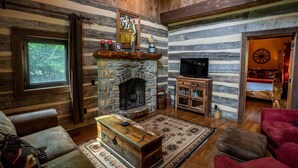 A rustic cabin interior with a stone fireplace, wooden furniture, a television, and a large window.