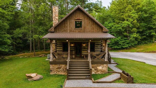 A rustic cabin with a stone chimney, a porch with steps, and a view of the surrounding forest.
