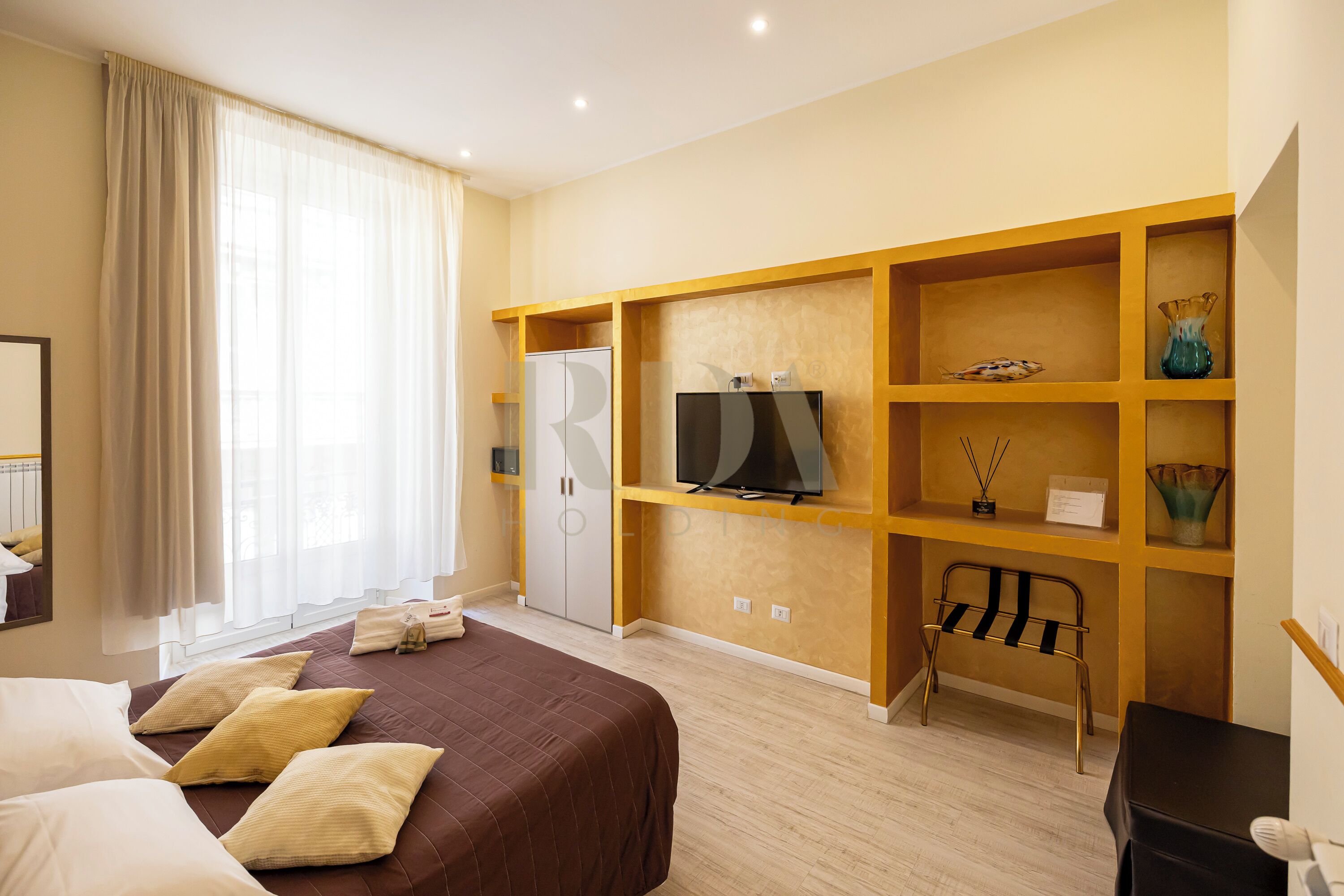 A hotel room with a bed, a TV, a wardrobe, and a shelf with decorative items.