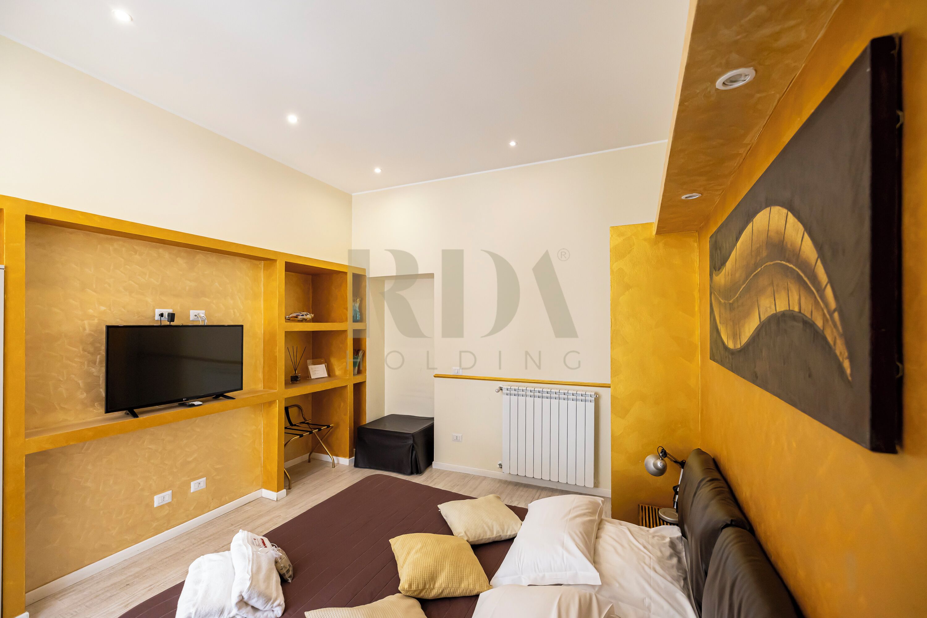 A modern hotel room with a flat-screen TV, wooden shelving, a bed with multiple pillows, a radiator, and a painting on the wall.