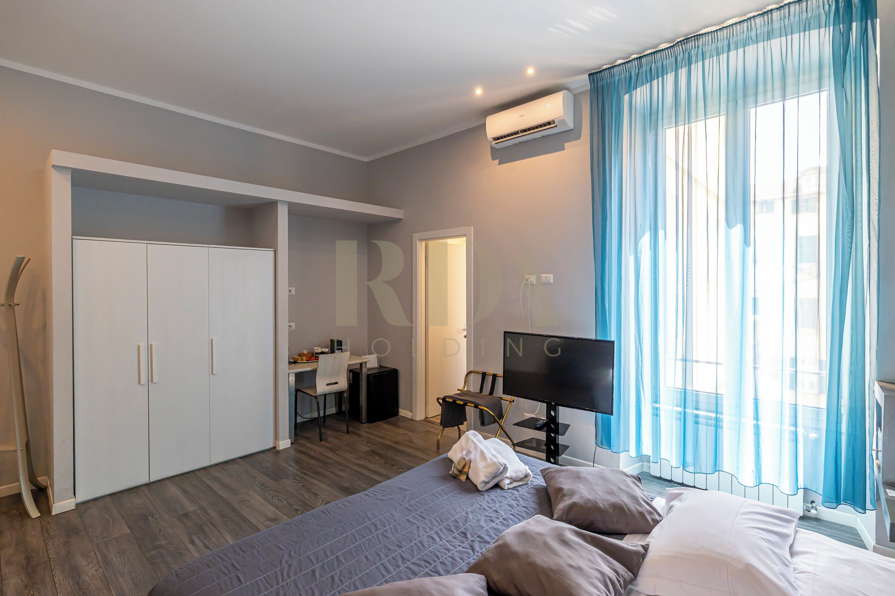 A bedroom with a bed, a desk, a chair, a wardrobe, and an air conditioning unit.