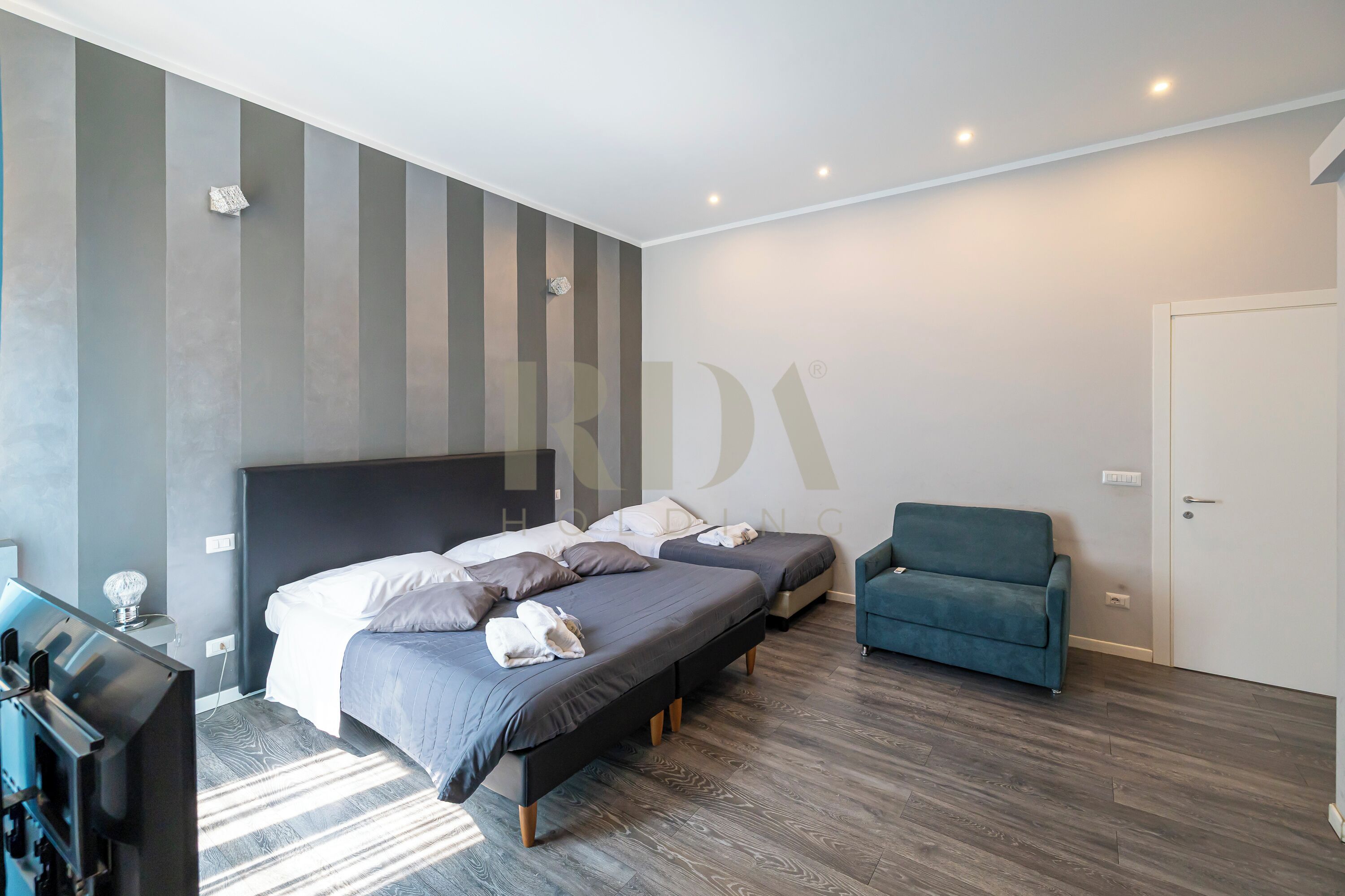 Una habitació d'hotel moderna amb dos llits, un sofà, una televisió i una làmpada fixada a la paret.