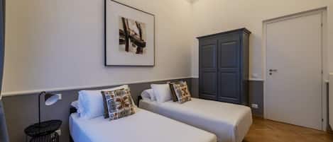 A hotel room with two beds, a wardrobe, a framed picture on the wall, and a small table with a lamp.