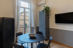 Ein Zimmer mit einem runden Tisch, Stühlen, einem Fernseher, einer Pflanze und Blick auf Gebäude durch ein Fenster.