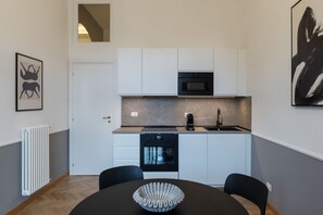 A compact kitchen with white cabinets, a black oven, and a microwave. There's a dining table with a decorative bowl and two chairs. - Castro Pretorio 25 - FHTR (Roma)