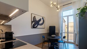 Une cuisine moderne équipée d’une machine à café, un coin repas avec une table et des chaises, une grande peinture abstraite et un balcon avec des rideaux.