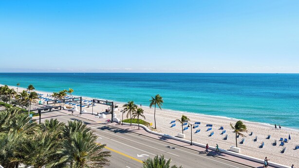 Ein Strand mit einer daran entlangführenden Straße, Palmen und wolkenlosem, blauem Himmel.