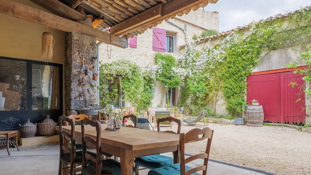 A rustic outdoor dining area with a wooden table and chairs, surrounded by stone walls and a red gate. - Guest Room “Côté Cour” with Large Shared Courtyard and Wi-Fi (Saint-Remèze)