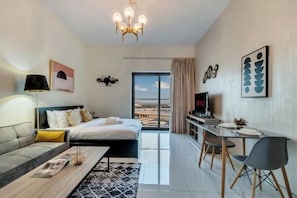 A modern hotel room with a bed, a desk, a TV, and a view of the city. - Elegant Studio with Balcony in Liwan – Ideal for Long Stays in Dubai (Dubai)