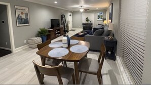 A modern living room with a grey sofa, a wooden dining table with placemats, and a television in the background. - Houston Home Near NRG Sleeps 6 (Houston)