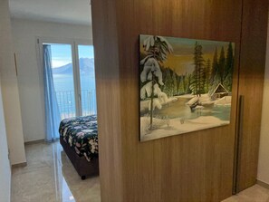 Une chambre à coucher avec un lit, un tableau représentant un paysage enneigé et une vue sur la mer depuis un balcon.