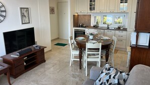 Une cuisine moderne avec des meubles blancs, un coin repas avec une table et des chaises en bois, et un salon avec un téléviseur et un canapé.