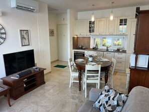 Une cuisine moderne avec des meubles blancs, un coin repas avec une table et des chaises en bois, et un salon avec un téléviseur et un canapé.