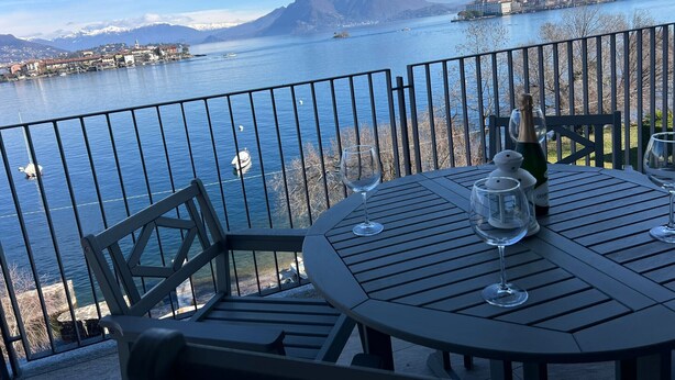 Une table avec des verres à vin et une bouteille, donnant sur un lac où l’on aperçoit des bateaux et une ville au loin.