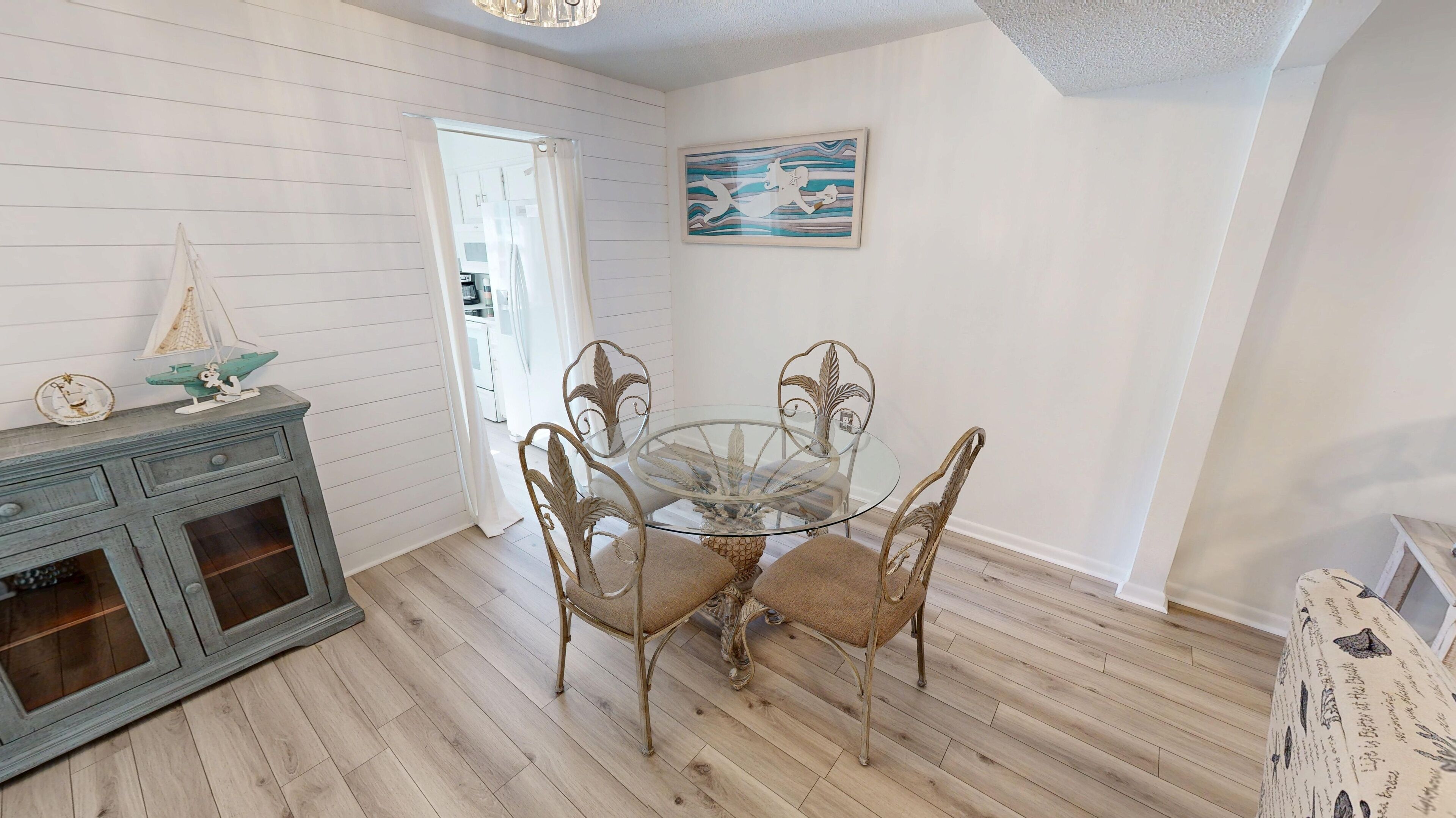 Zona pranzo con tavolo in vetro, quattro sedie, un mobile contenitore in legno con cassetti e una barca a vela decorativa.