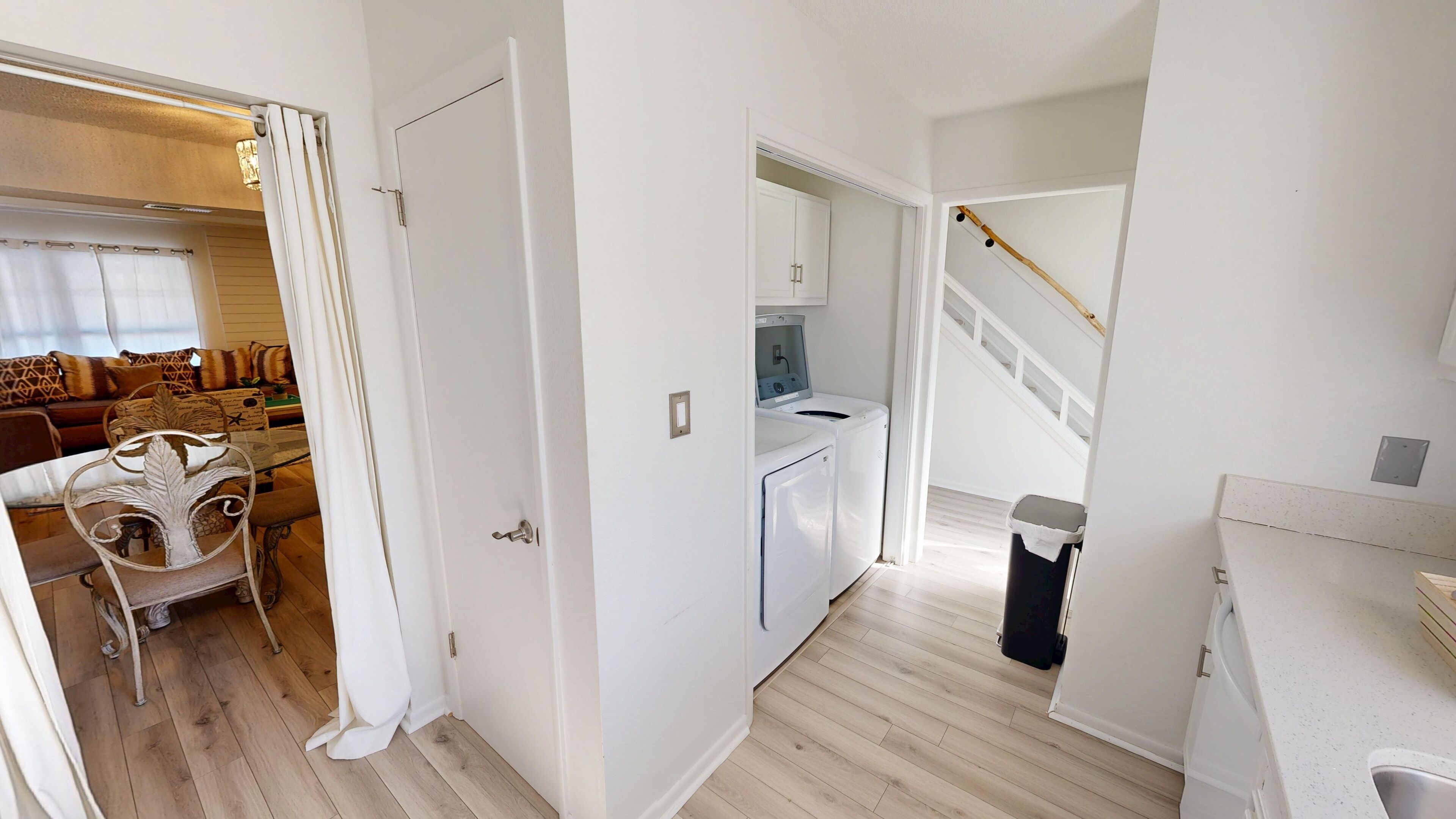 A compact, modern apartment with a living area featuring a sofa and a dining table, a laundry room with a washing machine and dryer, and a staircase leading to an upper floor.