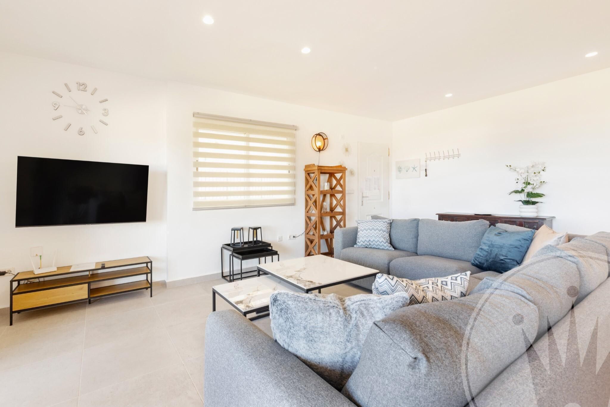 Un salon moderne avec un canapé gris, un téléviseur à écran plat, une table d’appoint en bois et une horloge murale décorative.