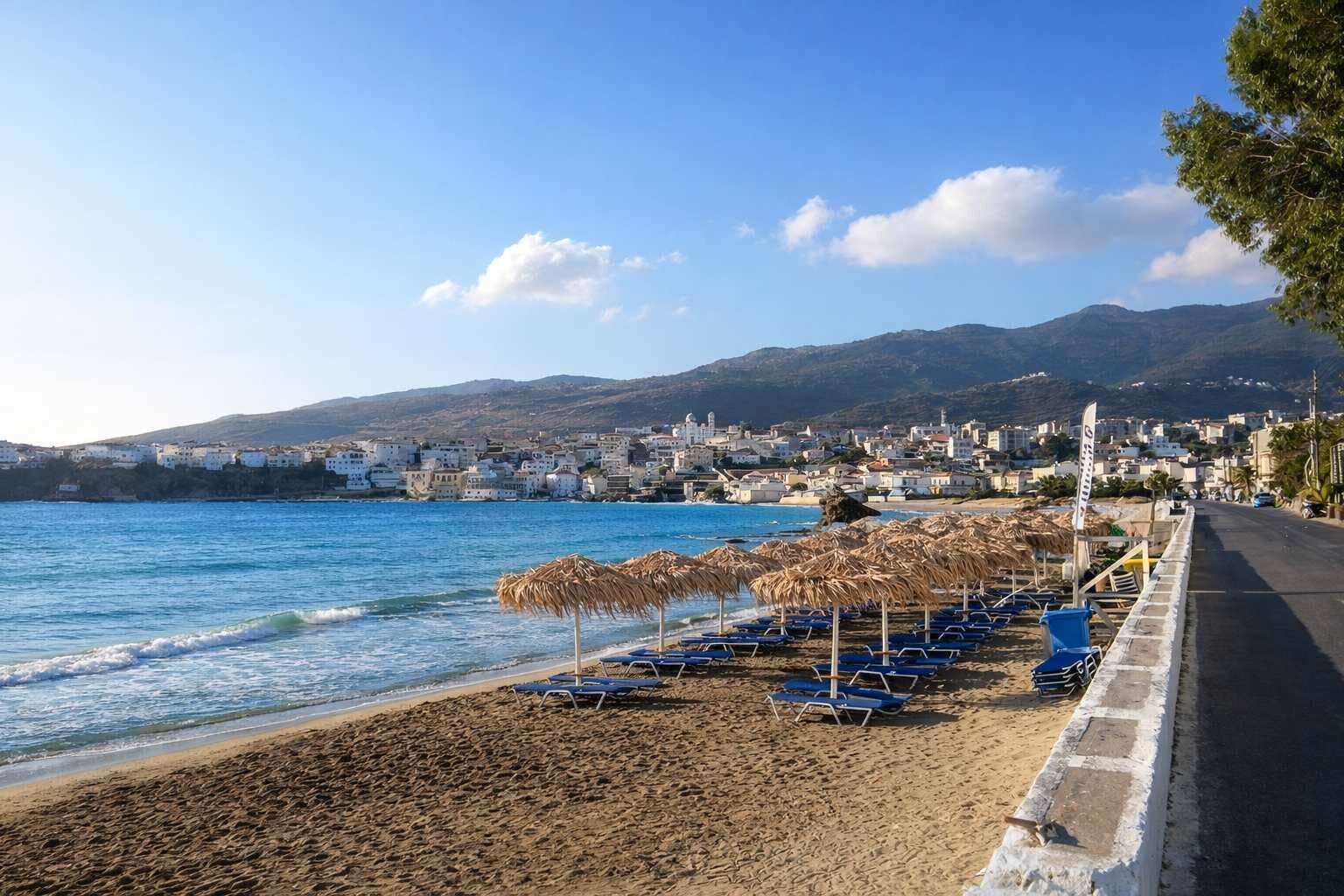 Una playa con sombrillas y sillas de descanso, un mar azul cristalino y un pueblo al fondo.