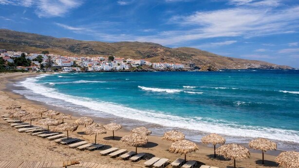 Una spiaggia con ombrelloni di paglia e lettini, un mare cristallino e una cittadina sullo sfondo.