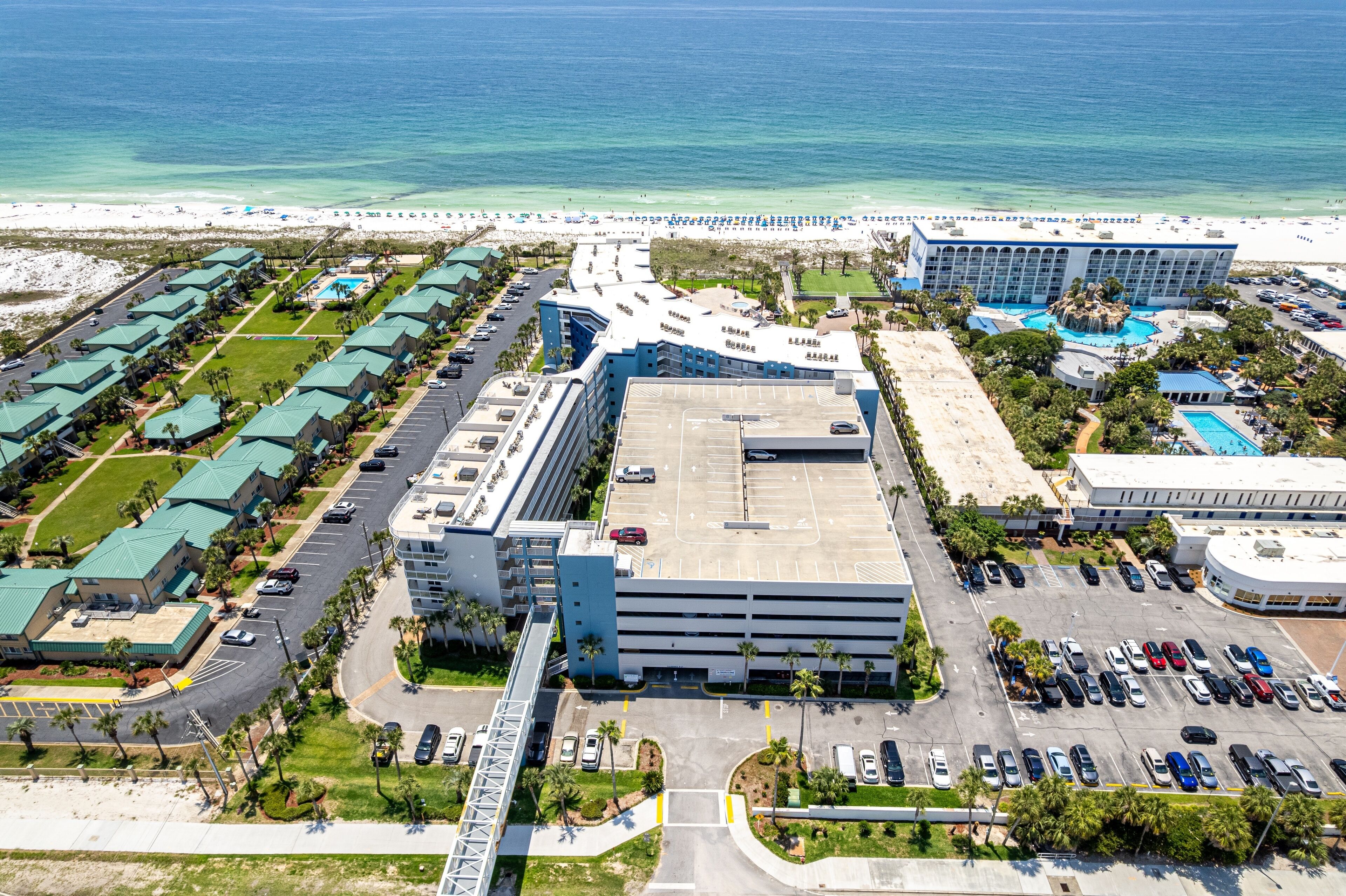 An aerial view of a hotel with multiple buildings, a parking lot, and a beach in the background.