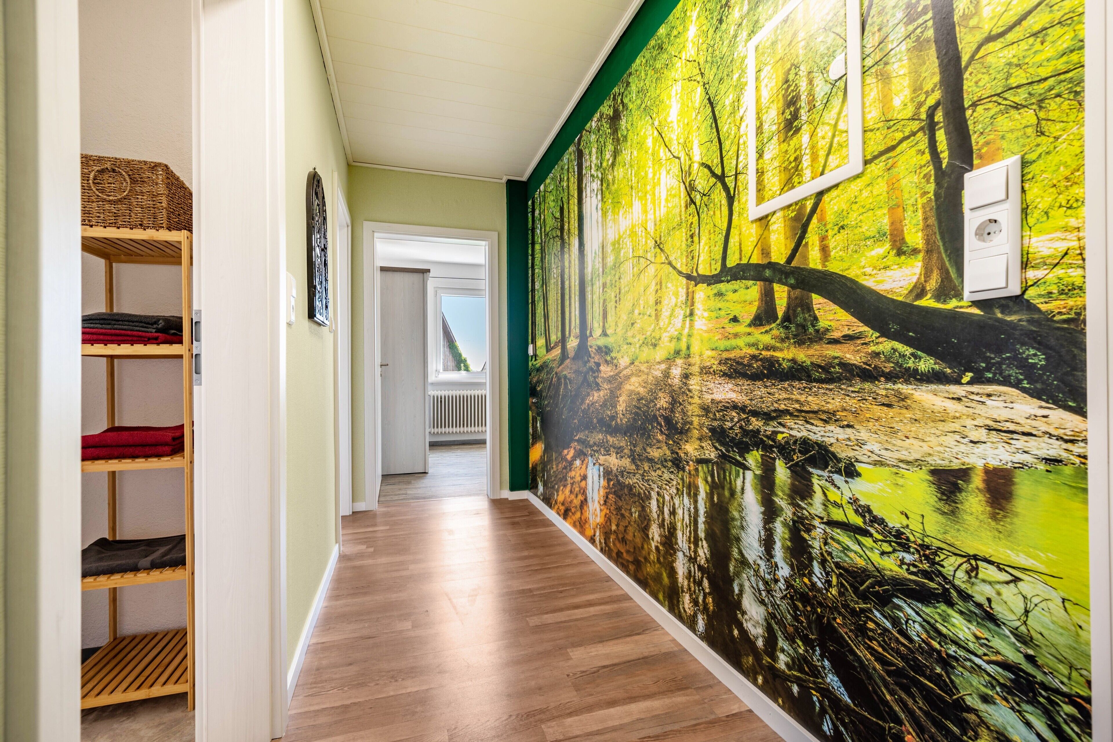 Un couloir doté d’un plancher en bois, d’une étagère murale où sont pliées des couvertures, et d’une grande fresque représentant une forêt.