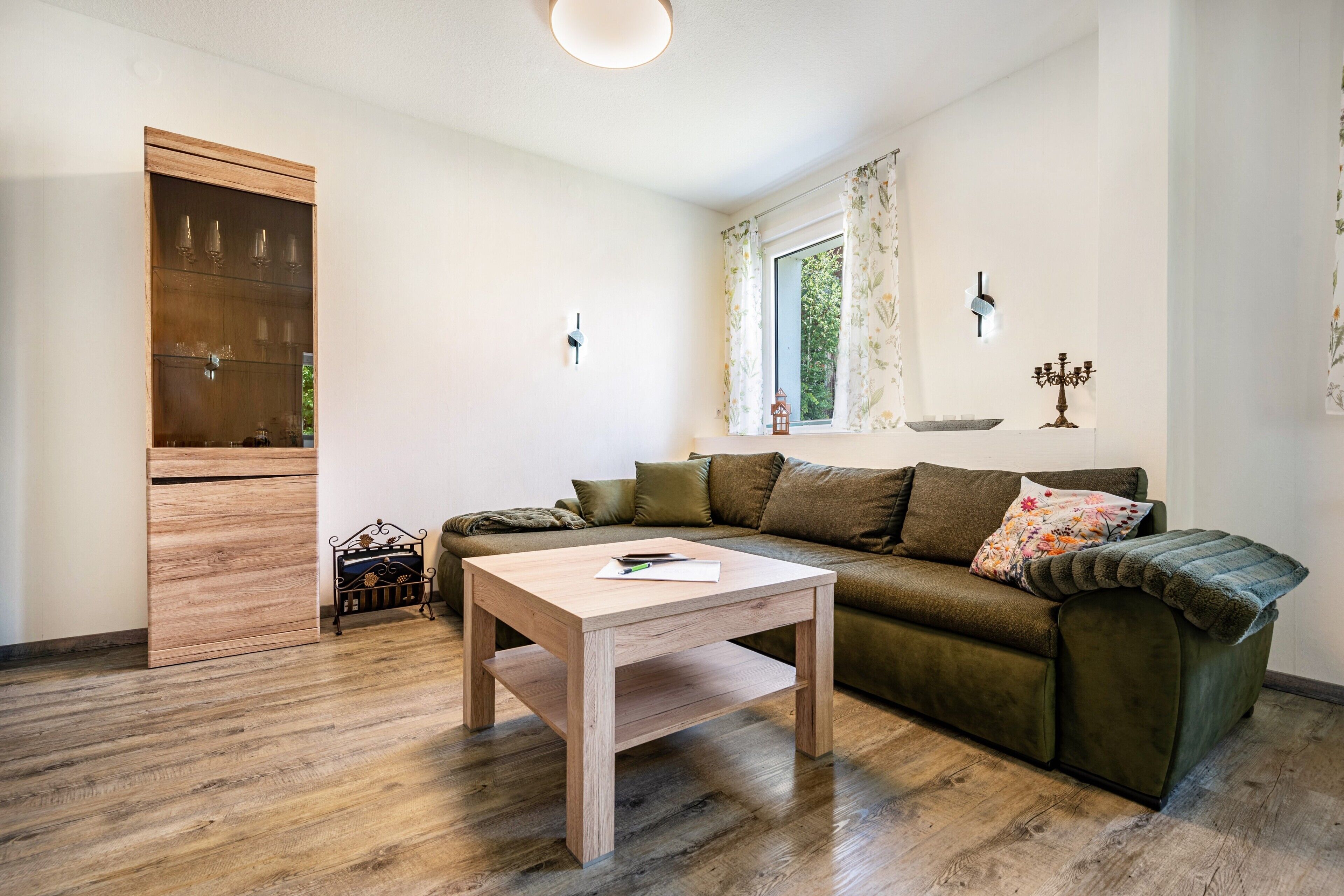 Ein Wohnzimmer mit Holzboden, einem grünen Sofa, einem Couchtisch aus Holz, einem Schrank und einem Fenster mit Vorhängen.