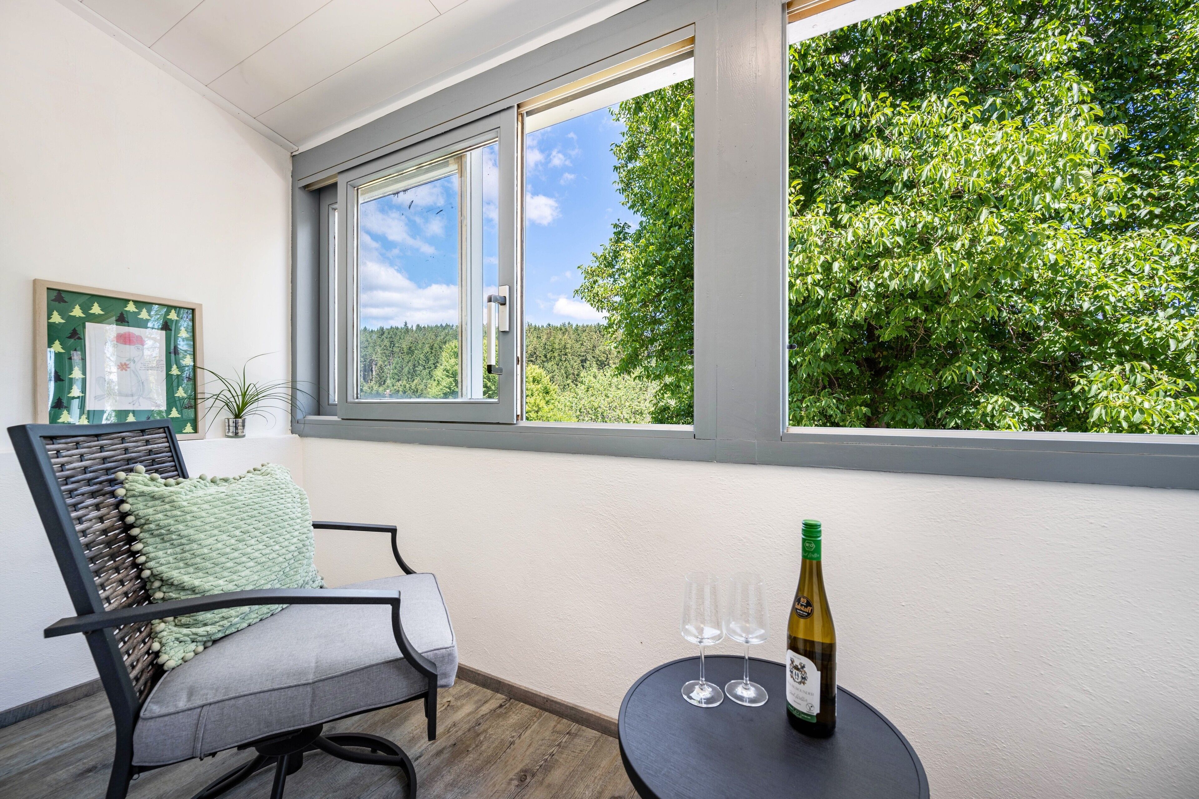 Et værelse med et vindue der udover en skov, en stol med pude og et bord med en flaske og glas.