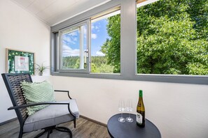 A room with a window overlooking a forest, a chair with a cushion, and a table with a bottle and glasses. - Apartment 'Ferienwohnung Zum Schwarzwaldfrosch' with Balcony and Wi-Fi (Freudenstadt)