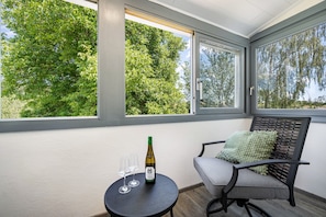 A room with a view of trees through large windows, a table with a bottle and glasses, and a chair with a cushion. - Apartment 'Ferienwohnung Zum Schwarzwaldfrosch' with Balcony and Wi-Fi (Freudenstadt)
