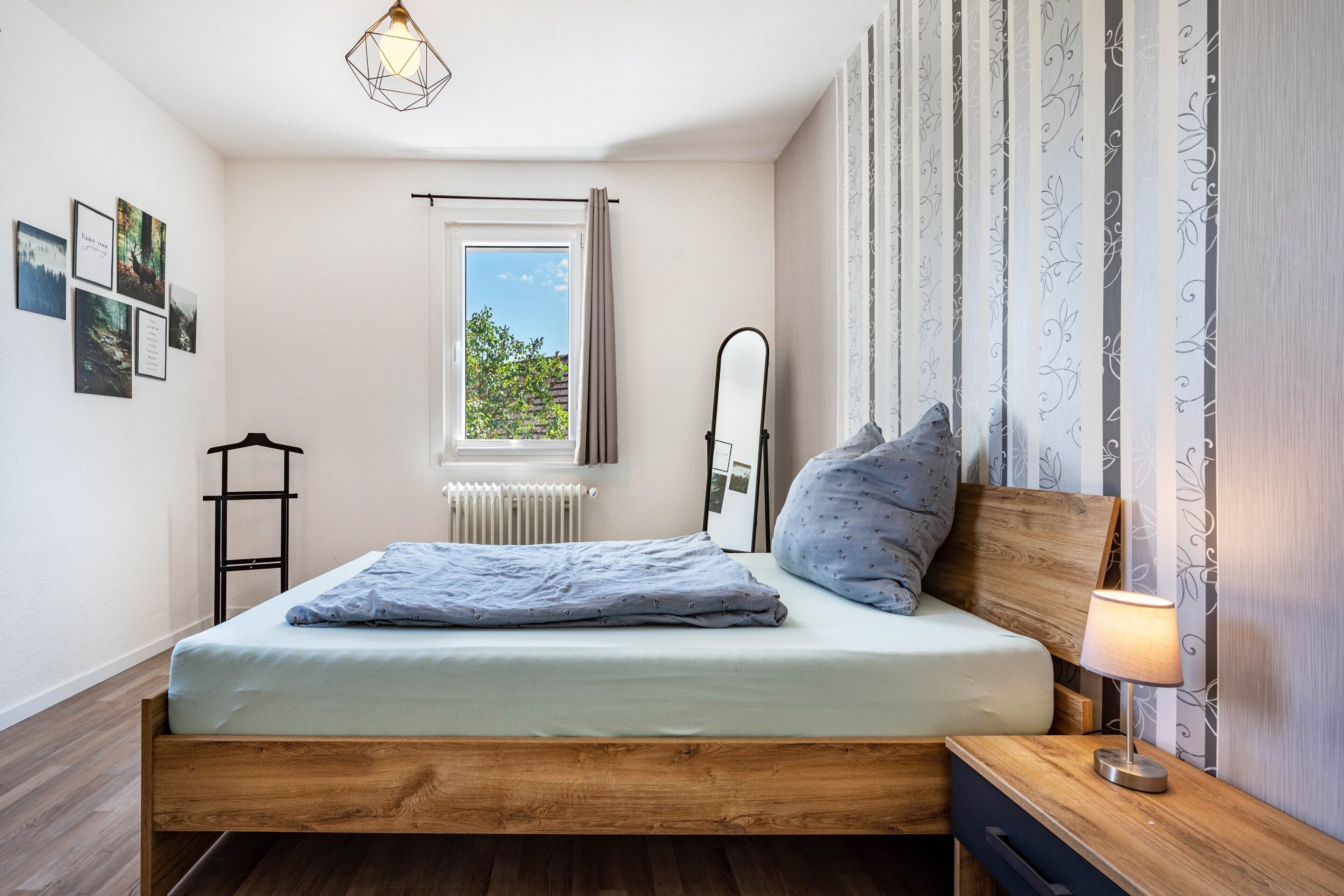 A bedroom with a wooden bed, a bedside table with a lamp, a radiator, a window with curtains, and framed pictures on the wall.