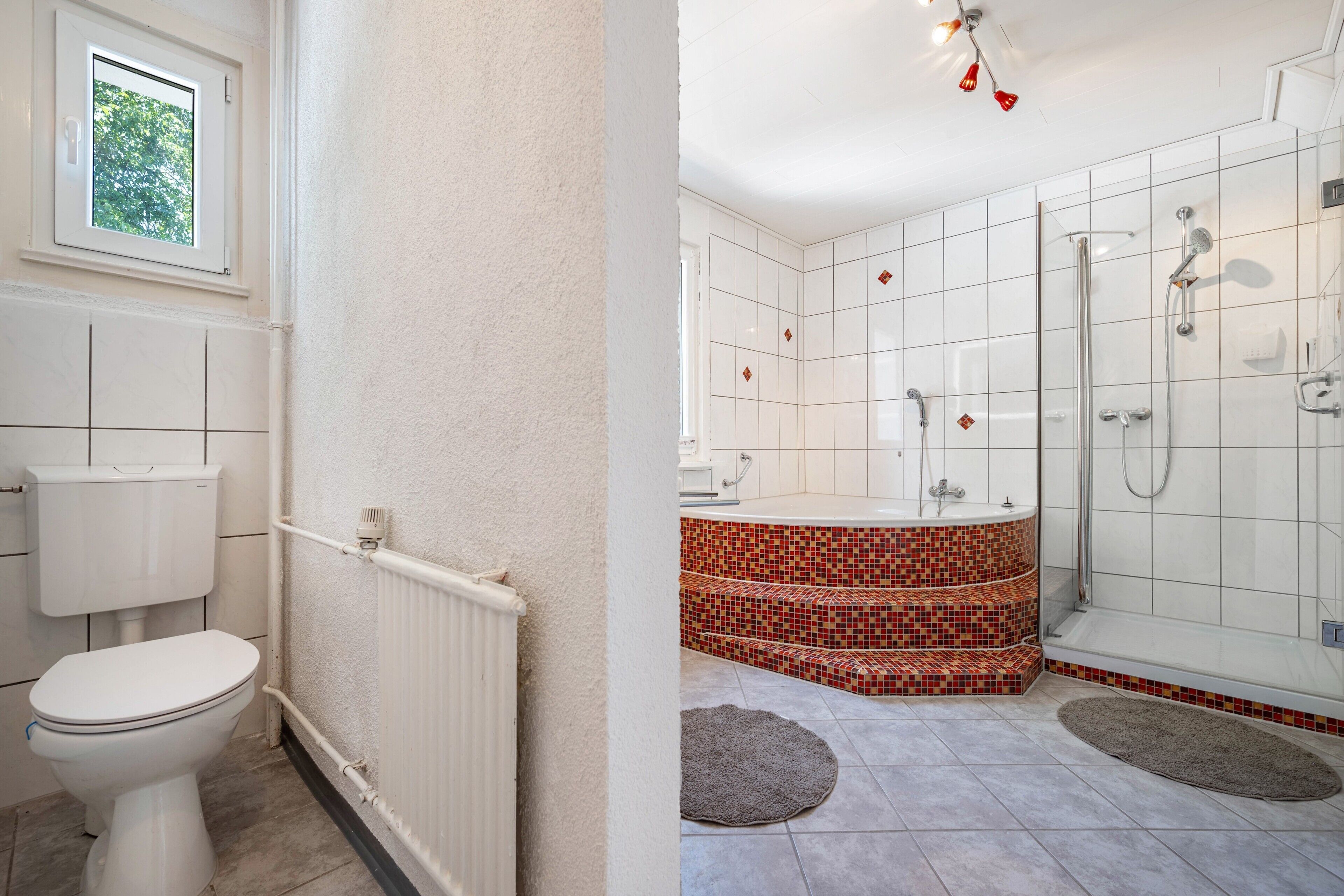 A bathroom with a toilet, a radiator, a shower with a glass door, and a window with a view of trees.