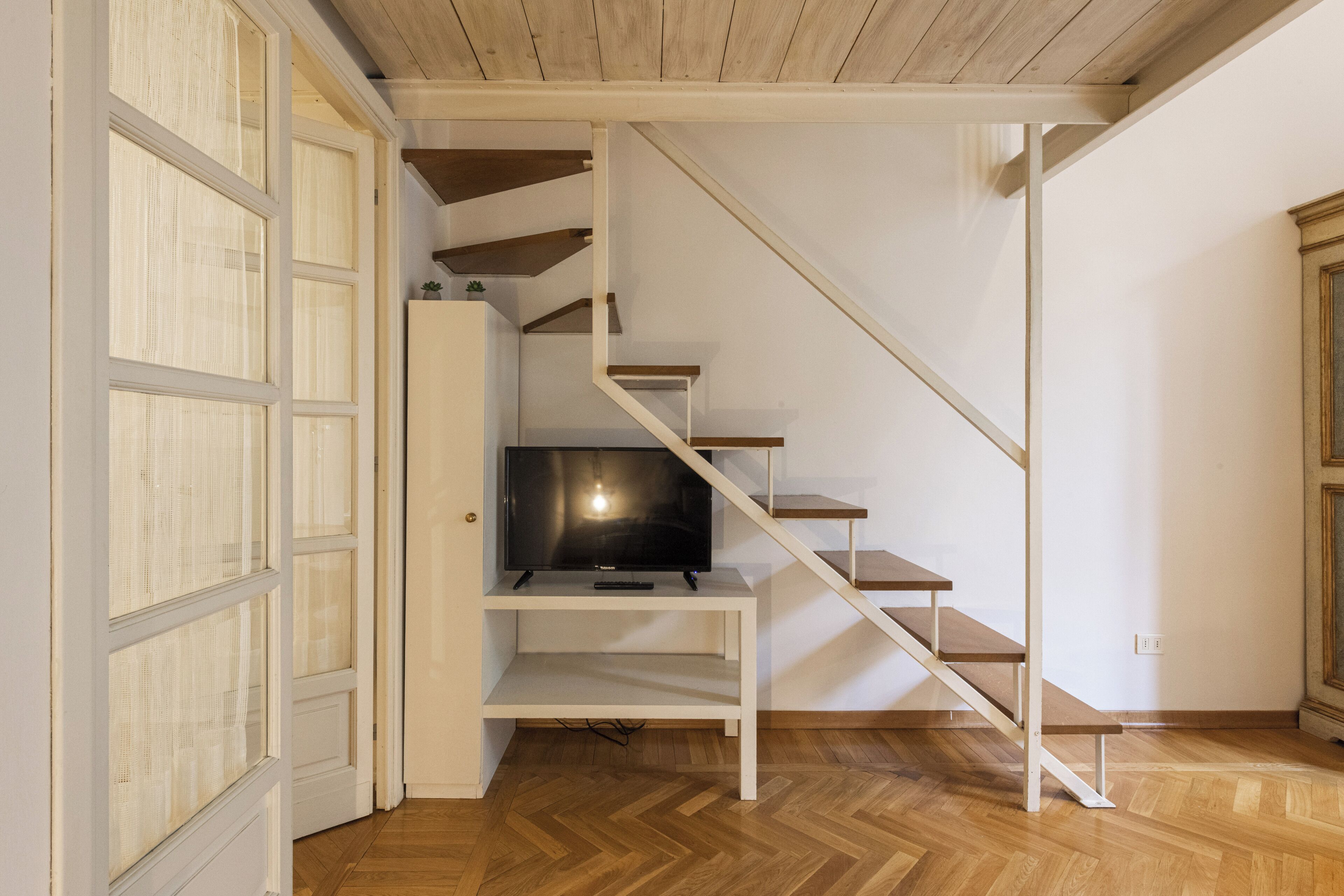 A staircase with a television on a shelf, wooden flooring, and a white door with glass panels.
